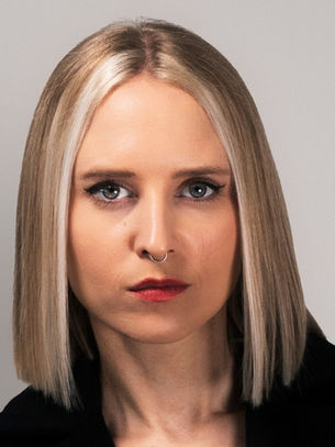 Portrait of a serious blonde woman with a septum ring in a black top against a plain gray background.