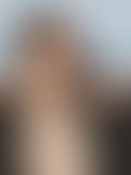 Man wearing sunglasses adjusts coat on a sunny day, standing with a cityscape backdrop. Casual attire; relaxed expression.