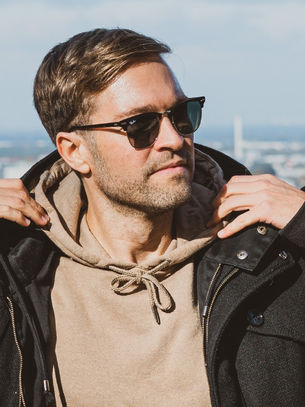 Man wearing sunglasses adjusts coat on a sunny day, standing with a cityscape backdrop. Casual attire; relaxed expression.