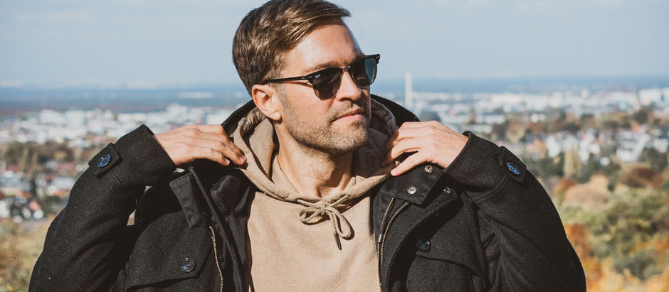 Man wearing sunglasses adjusts coat on a sunny day, standing with a cityscape backdrop. Casual attire; relaxed expression.