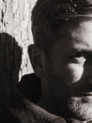 Man with light stubble looks contemplative, leaning against a tree in black and white. Sunlight casts shadows, creating a dramatic effect.