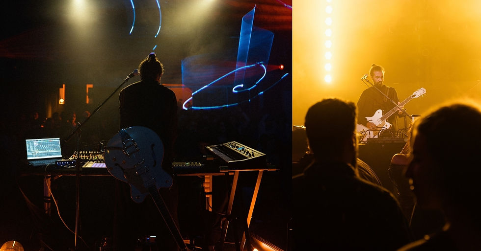 Live concert with silhouetted performer at electronics on left and guitarist on right, bathed in warm yellow stage lights, crowd in front