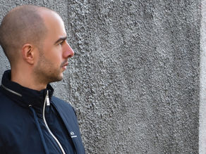 Bald man in a dark jacket stands in profile against a textured gray wall, looking right with a neutral expression.