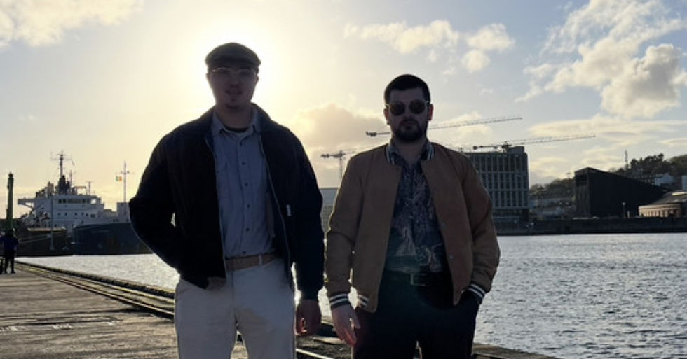 Two men stand on a waterfront dock at sunset, with ships, cranes, and city buildings in the background.