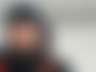 Man in a snowy landscape, wearing a dark jacket and beanie, with frost on his beard. Overcast sky, icy terrain, and a red backpack strap.