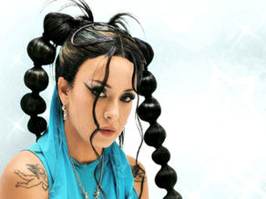 Portrait of a woman with dramatic bubble braids, blue top, tattoos and piercings against a pale glowing background
