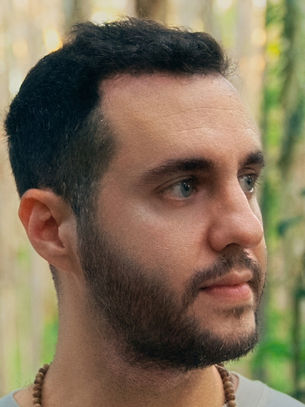 Man with short hair and beard looks to the side in a forest, wearing a beaded necklace. Background is lush with trees and greenery.