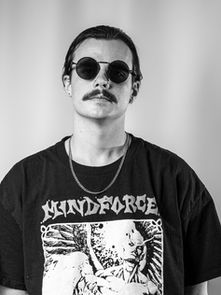 Person with sunglasses and a mustache wearing a "Mindforce" T-shirt stands in front of a plain background, exuding a calm demeanor.