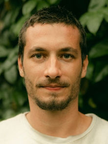A man with short hair and a beard stands in front of lush green leaves. He wears a neutral expression and a white shirt.