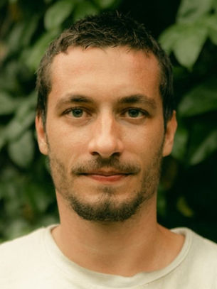A man with short hair and a beard stands in front of lush green leaves. He wears a neutral expression and a white shirt.