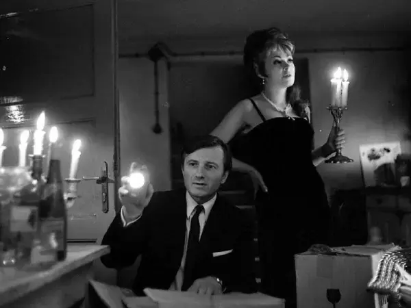 A woman stands behind a seated man in a candlelit house.