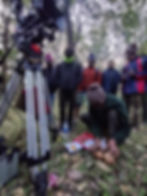 A camera crew wearing hats outside breaks a coconut next to the camera.