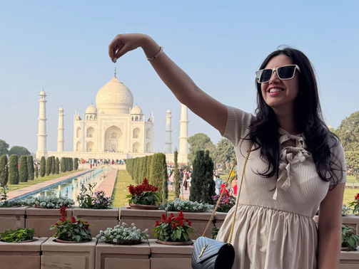A woman playfully poses as if she is pinching the top of the Taj Mahal in Agra, India, with a smile and sunglasses under a clear blue sky.
