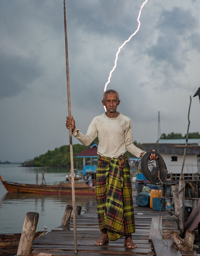 A portrait of an Indonesian fisherman on a jetty with a strike of lighting behind him.
