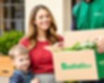 A woman and child stand in front of their house receiving a delivery of vegetables in box. The box is labeled BushelBox.com.