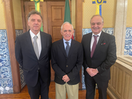 Foto em plano médio de três homens em ambiente oficial com paredes de azulejos portugueses azuis e brancos. À esquerda, Antônio Guedes de terno azul-marinho e gravata cinza; ao centro, Raimundo Carreiro Silva de blazer escuro, camisa azul e calça bege; à direita, Nelson Rocha, diretor da CISBRA, de terno escuro e gravata bordô. Todos estão em pé, posicionados lado a lado, sorrindo ou em postura formal.