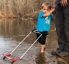 2-year-old blind boy stands in on the shore in shallow water wearing his belt cane, holds his dad's hand.