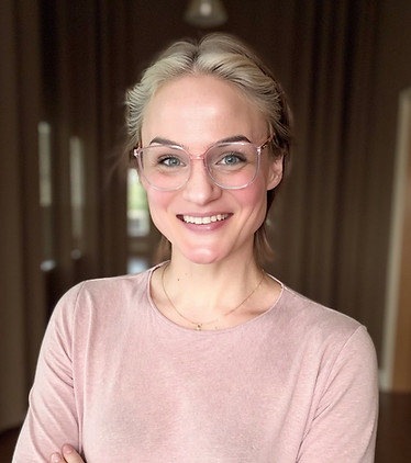 Smiling female clinical supervisor and spiritual guide wearing glasses and a light pink top, standing confidently in a warmly lit hallway.