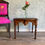 Thumbnail: front view of wood, twisted leg table with a vase on top with one flower. A brightly painted chair sitting next to it is partially showing.