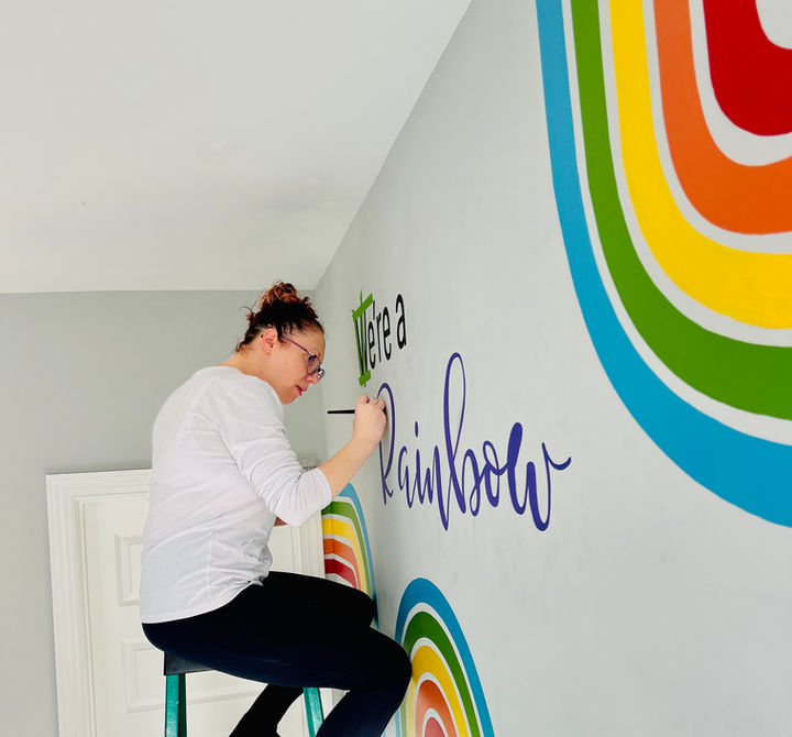 Artist painting a custom rainbow mural with hand-lettered text in a kids bedroom