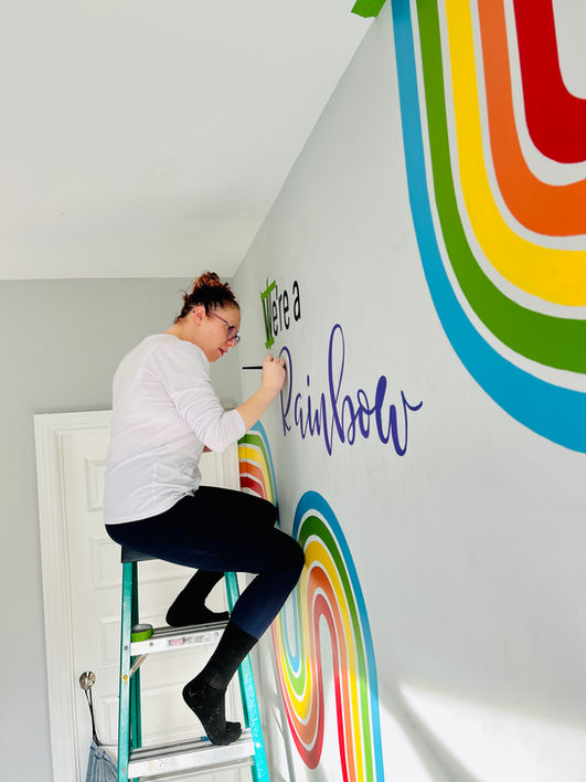 Artist painting a custom rainbow mural with hand-lettered text in a kids bedroom