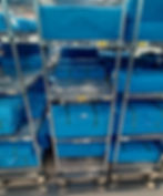 Blue steile packages are stacked on metal shelves with wheels in a sterile processing area. Shelves are labeled with yellow tags. Hospital setting.
