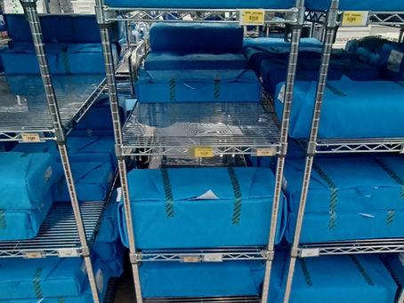 Blue steile packages are stacked on metal shelves with wheels in a sterile processing area. Shelves are labeled with yellow tags. Hospital setting.