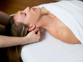 Woman lying face up on a massage table under a white sheet and blanket while a massage therapist gently massages her neck, illustrating a calm and relaxing spa environment for the blog post “What to Expect During Your First Massage.”