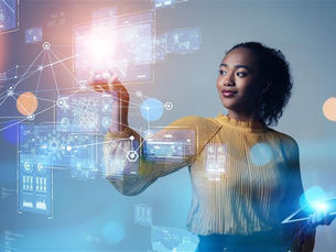 Woman in a yellow sweater uses a tablet and reaches toward glowing holographic data screens in a blue tech setting.