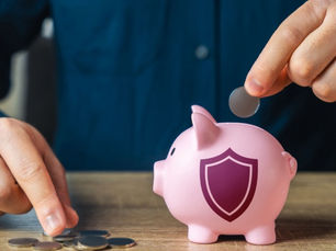 Hands place coins into a pink piggy bank with a shield icon on a wooden table, suggesting savings and security.