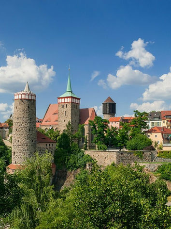 Historische Stadtansicht mit Türmen, Gebäuden und Bäumen unter blauem Himmel mit Wolken.