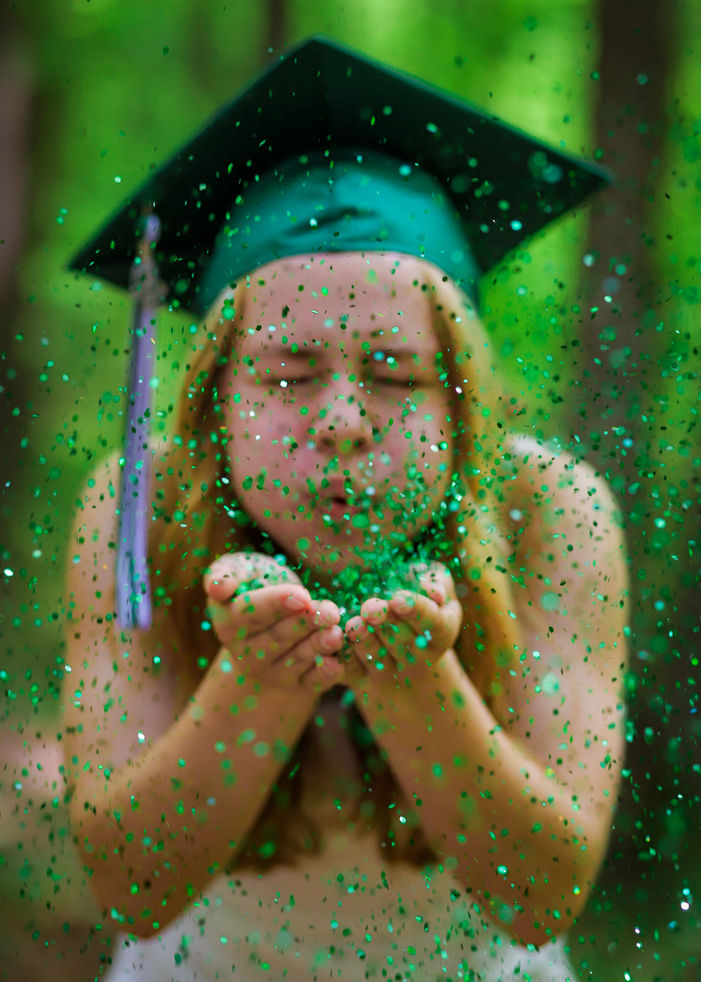 Young girl in graduation cap blowing glitter outdoors for celebration.