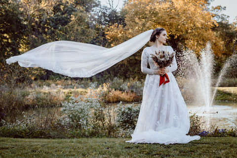 Bride in white dress, veil flying in the wind, near fountain with trees. LoveNikily Photos