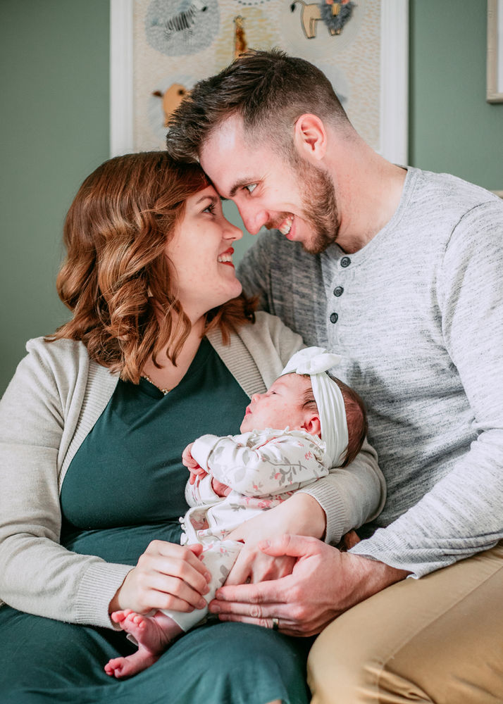 Parents lovingly gaze at newborn baby wearing a white bow. LoveNikily Photos