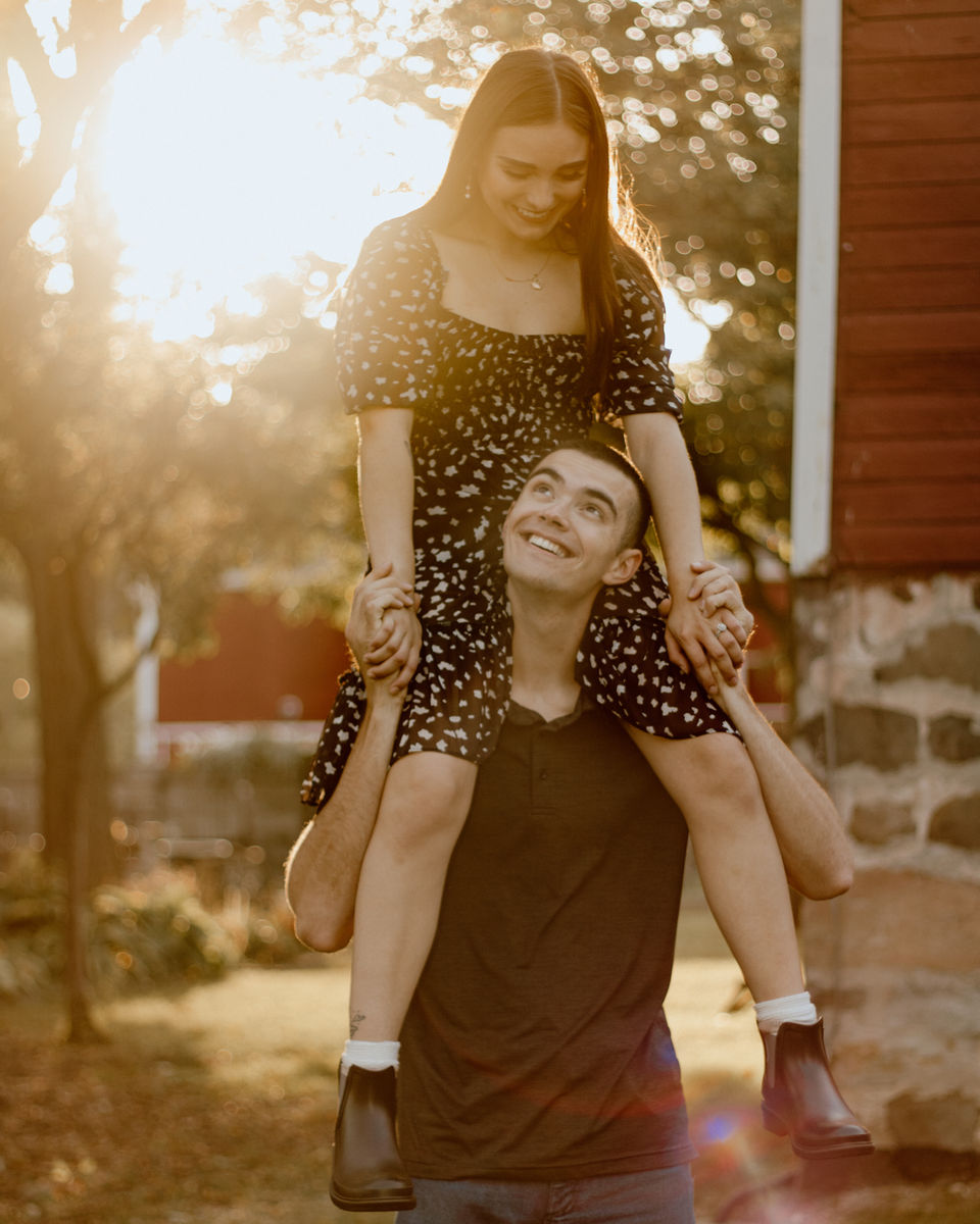 Young man carrying a woman on his shoulders, smiling in the sunlight. LoveNikily Photos