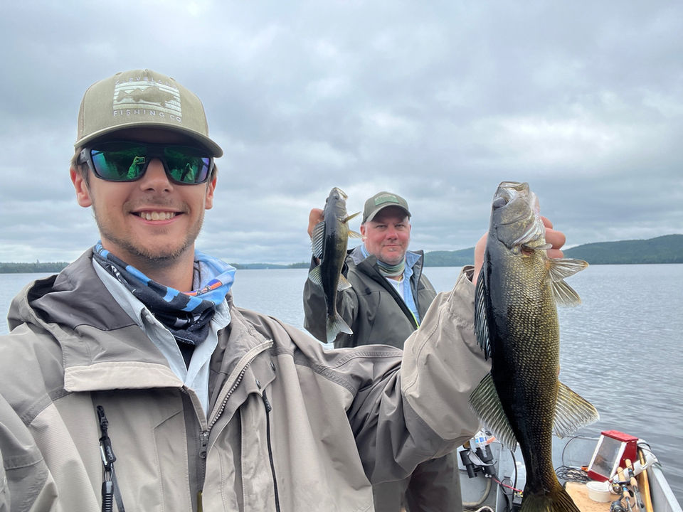 Two men standing side by side each holding nice walleye. The background is cloudy skies and calm lake water.