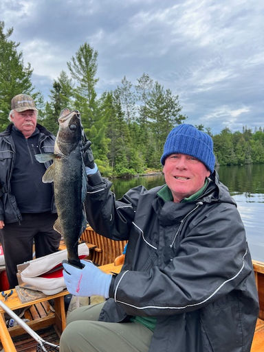 On a cool, cloudy 2 men sit in a WC-14 fishing boat. 1 man is holding a nice walleye.