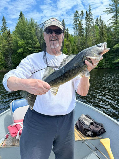 A man standing in a Lund wc-14 fishing boat holding a trophy walleye. The boat is anchored near shore.