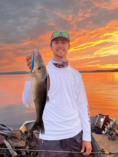 Man in a white t-shirt standing in a boat holding a nice walley. The backdrop is a stunning golden sunset.