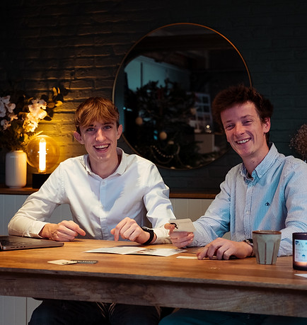 Emile (links) en Richard (rechts) van RE-TINK zitten aan een houten tafel in een sfeervolle ruimte. Emile, gekleed in een lic