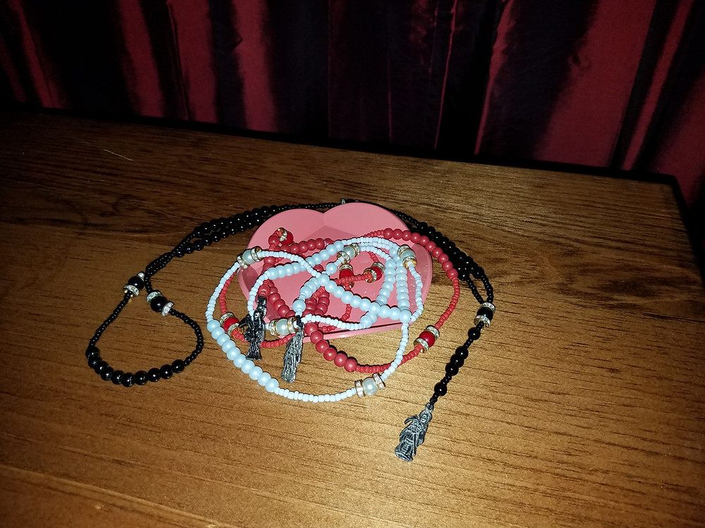 Three Santa Muerte rosaries, one white, one red, and one black, in a lotus dish on a table backed by a crimson drapery.