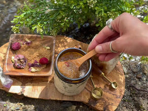 A jar of brown sugar scrub and a bowl of brown sugar with flower petals on a wooden slab with crystals and measuring spoons on the shore of a creek surrounded by plants and river rocks.