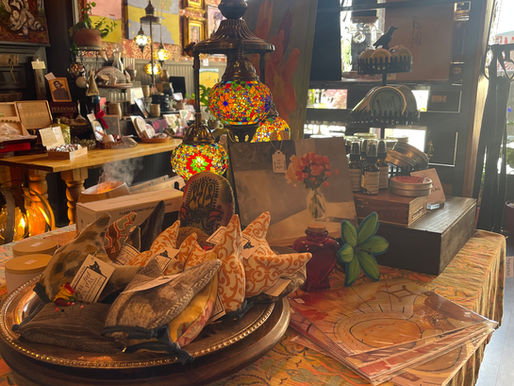 Display table inside The Wartrace Emporium featuring colorful lamps, art prints, candles, and handmade gifts, with patterned wallpaper and framed artwork in the background.
