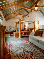 Spacious cabin suite with exposed wood beams, a log four-poster bed, and a patterned rug under a vaulted metal ceiling.