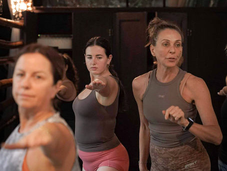 Women in workout attire practice yoga in a dimly lit studio. They extend arms in focus; background shows shelves and soft lighting.