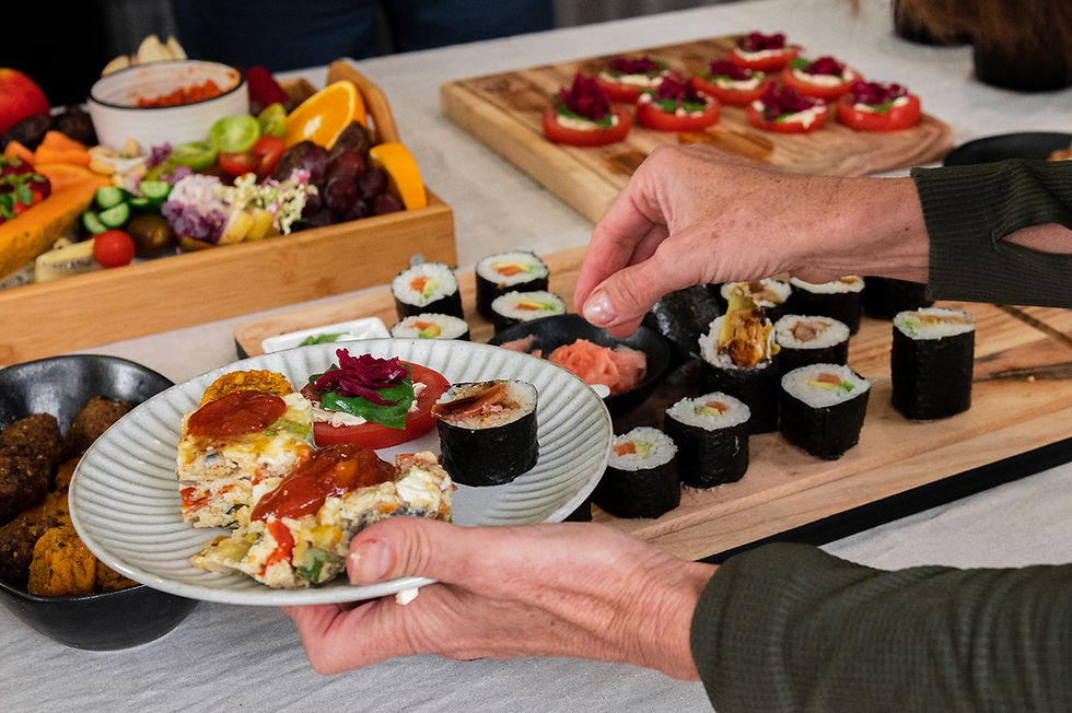 Hands holding a plate with sushi, salad, and quiche. A table displays assorted sushi, veggies, and fruits on wooden trays. Vibrant and inviting.