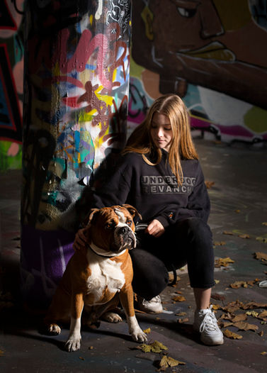 Junge Frau in der Hocke vor einer Graffiti Säule. Vor ihr sitzt ein Continental Bulldog. Sie trägt einen schwarzen oversized Hoodie mit einem gummierten, schwarzen Druck mit dem brand name Underdog Revanche