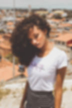 Junge Frau mit lockigen Haaren trägt weißes RSPCT T-Shirt von Underdog Revanche, urbanes Setting mit Häusern im Hintergrund