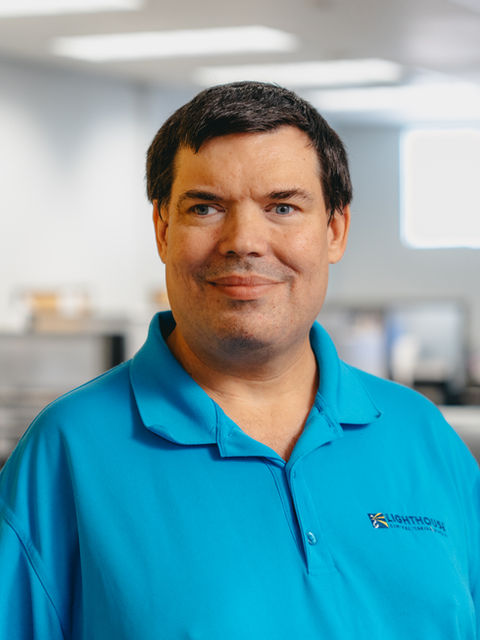 A man in a polo shirt with a Lighthouse logo smiling in an office with desks and computers.