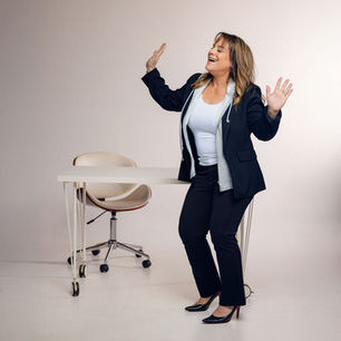 Woman in suit joyfully gesturing beside a modern desk and chair, set against a plain, light background. Mood is lively and upbeat.